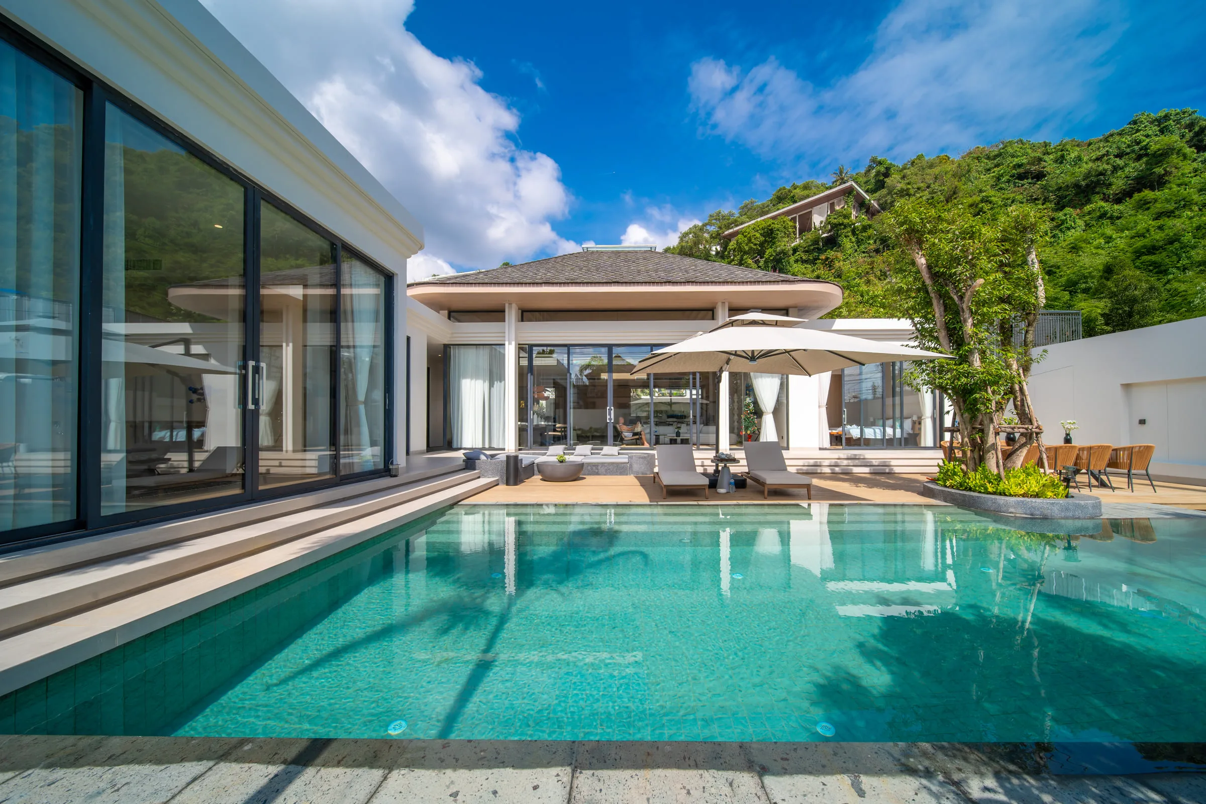 Villa exterior seen across the swimming pool, with a wooded hillside behind the roofline
